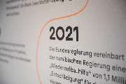 Bande für Gestaltung - Ausstellungselement mit der Jahreszahl 2021, das die politische und gesellschaftliche Aufarbeitung kolonialer Geschichte thematisiert. Eine leuchtend orangefarbene Linie führt durch den Text und symbolisiert den historischen Zusamme