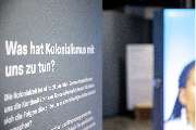 Bande für Gestaltung - Textwand der Ausstellung mit der Frage „Was hat Kolonialismus mit uns zu tun?“ zu sehen. Im Hintergrund erscheinen großformatige Porträts, die den Bezug zwischen Geschichte und Gegenwart sichtbar machen.