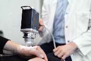 Dermatologikum Hamburg - Ein:e Ärzt:in untersucht die Haut einer Patient:in an der Hand mithilfe eines modernen Videodermatoskops. Das Gerät ist deutlich größer als ein klassisches Handdermatoskop und ermöglicht eine hochauflösende Bildübertragung zur exa