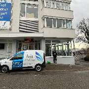 Fein Gebäude-Service - Ein weißes Fahrzeug mit Firmenlogo von Fein Gebäude-Service steht vor einem mehrstöckigen Gebäude, während eine Reinigungskraft die Fenster säubert.