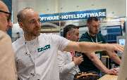 Gastronovi GmbH - Gastronovi Messestand: Mitarbeiter in weißem Shirt erklärt die Software live am Bildschirm. Besucher verfolgen die Demo der Gastro-Software. Banner im Hintergrund sichtbar.