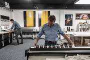 IB Gosch - fineArt prints & Einrahmungen - Einrahmungsexperte Frank Reimer von der Firma IB Gosch an der Schneidemaschine mit Druckmustern auf Fine Art Papier. Im Hintergrund die Großformatdrucker von der Firma Epson neben Geschäftsinhaber Marius Gosch
