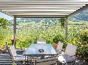 Mario Maierl GmbH München Markisen Lamellendach Sonnenschutz Jalousien Rollladen Rollo Reparatur - halb-transparente, weiße Standmarkise über einer Terrasse mit Glas-Tisch und Stühlen, inmitten der Natur.