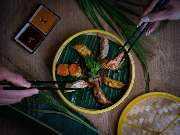 MAUI - Hawasian & Vietnamese Restaurant | München Ludwigpalais - Asiatische Spezialitätenplatte mit Gyoza, Shrimps und Gemüse, serviert auf Bambusteller mit Dip und dekorativem Palmblatt – zwei Personen greifen mit Stäbchen zu.