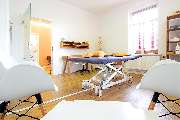 Physiotherapie Praxis Stefan Arnhold - Ein geräumiger Behandlungsraum mit einer zentral platzierten, höhenverstellbaren Therapieliege mit dunkelgrauem Bezug. Mehrere ergonomische, orangefarbene Polsterkissen liegen darauf. Der Raum hat helle Wände und ein